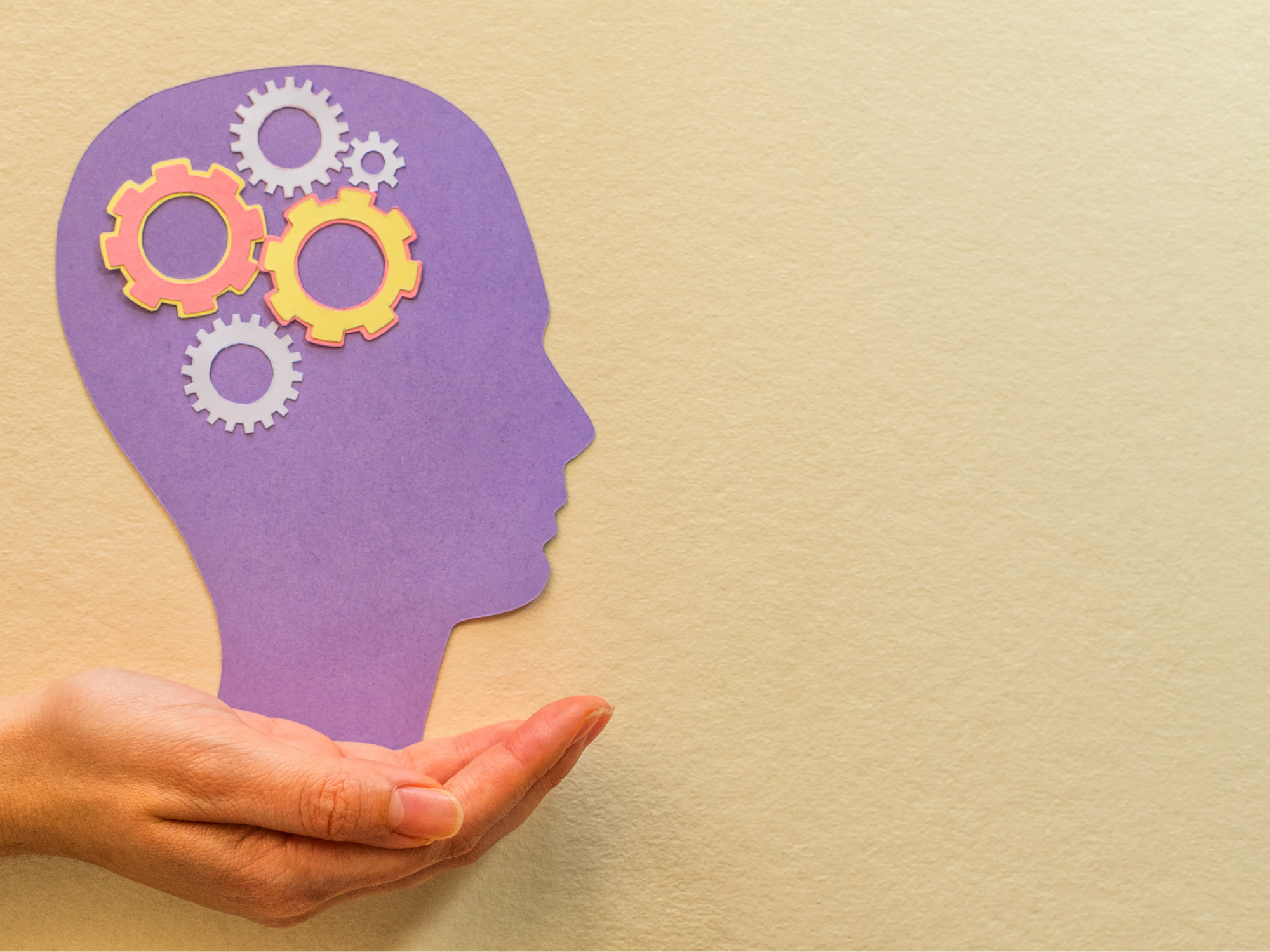 Hand holding a paper cut out of a purple head with cogs in the area of the head where the brain typically is.
